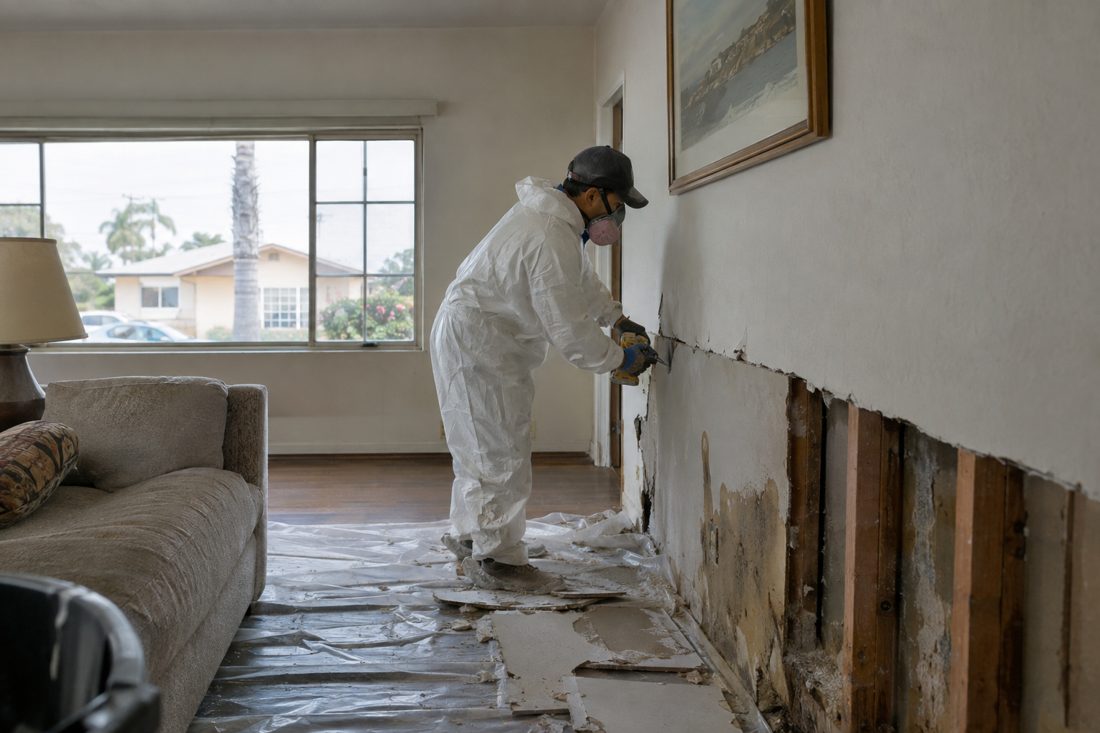 A technician in a tyvek suit cutting out wet drywall two feet up the wall in a living room at a 1950s stucco single-story bungalow in a Southern California neighborhood in Gardena, CA