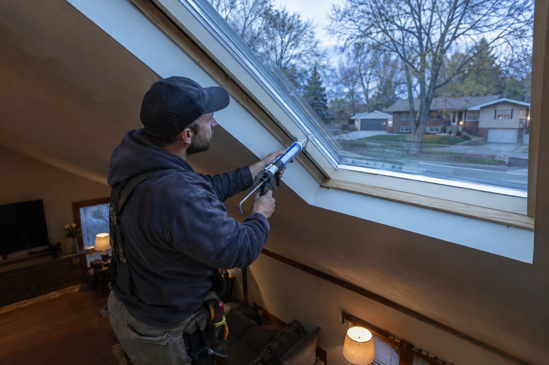 A roofer applying sealant around a skylight at a suburban Iowa split-level with oak trees in Hiawatha, IA