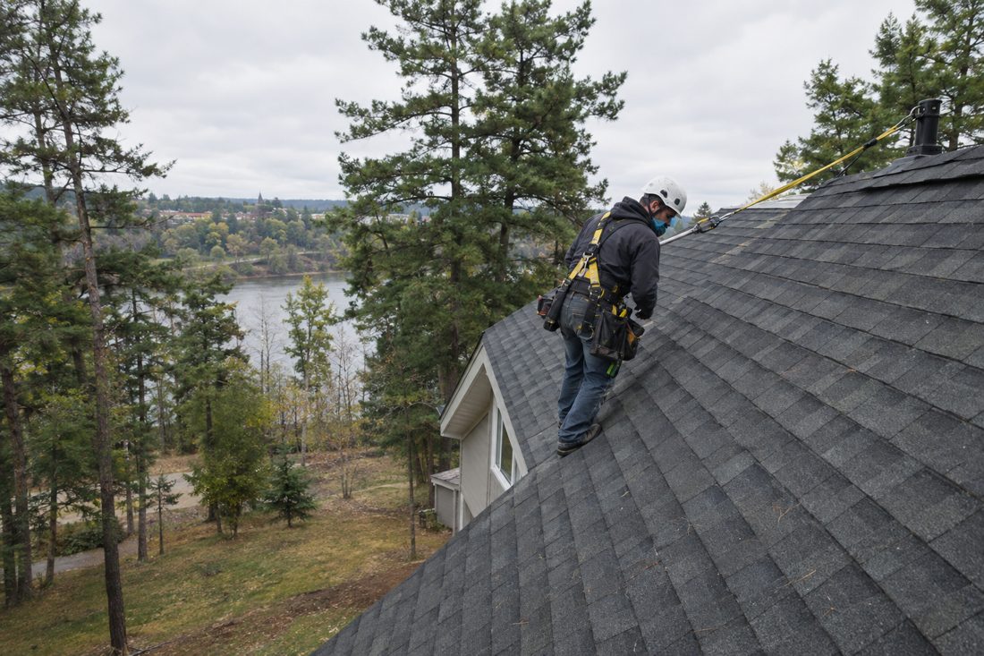 A crew member in a harness tied off to a roof anchor at a central Minnesota home with a steep roof and tall pines in Little Falls, MN